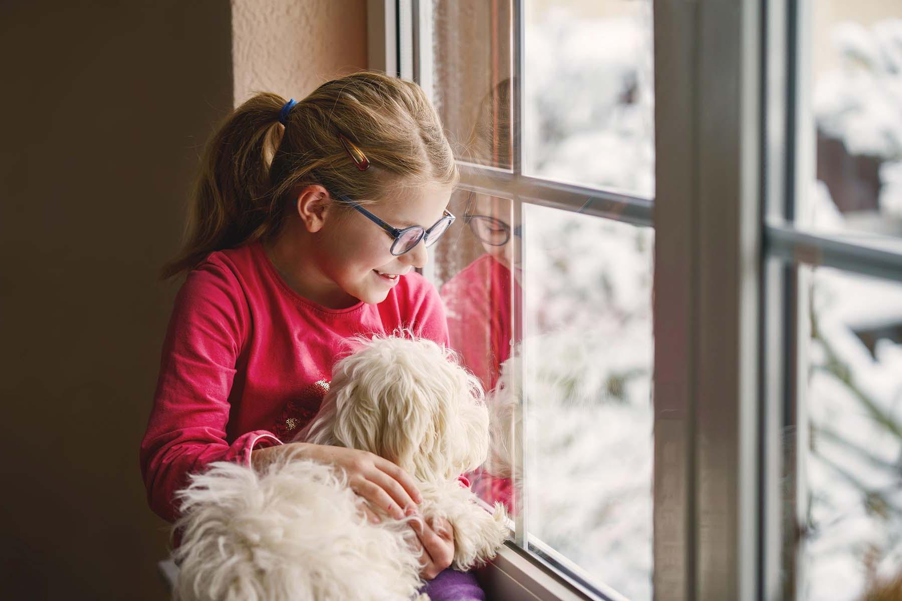 Girl with glasses holding a dog, looking out a snowy window.